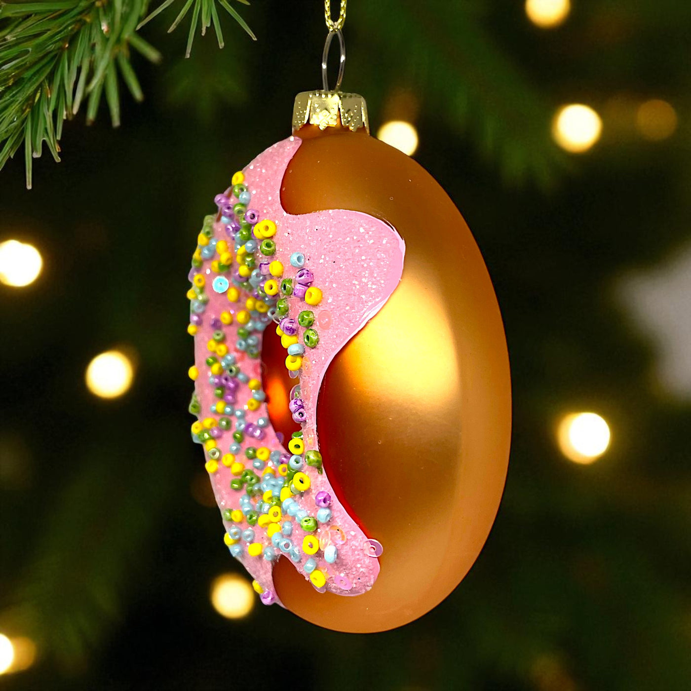 Decorative Christmas ornament shaped like a donut with pink icing and colorful sprinkles, hanging on a tree.