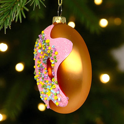 Decorative Christmas ornament shaped like a donut with pink icing and colorful sprinkles, hanging on a tree.
