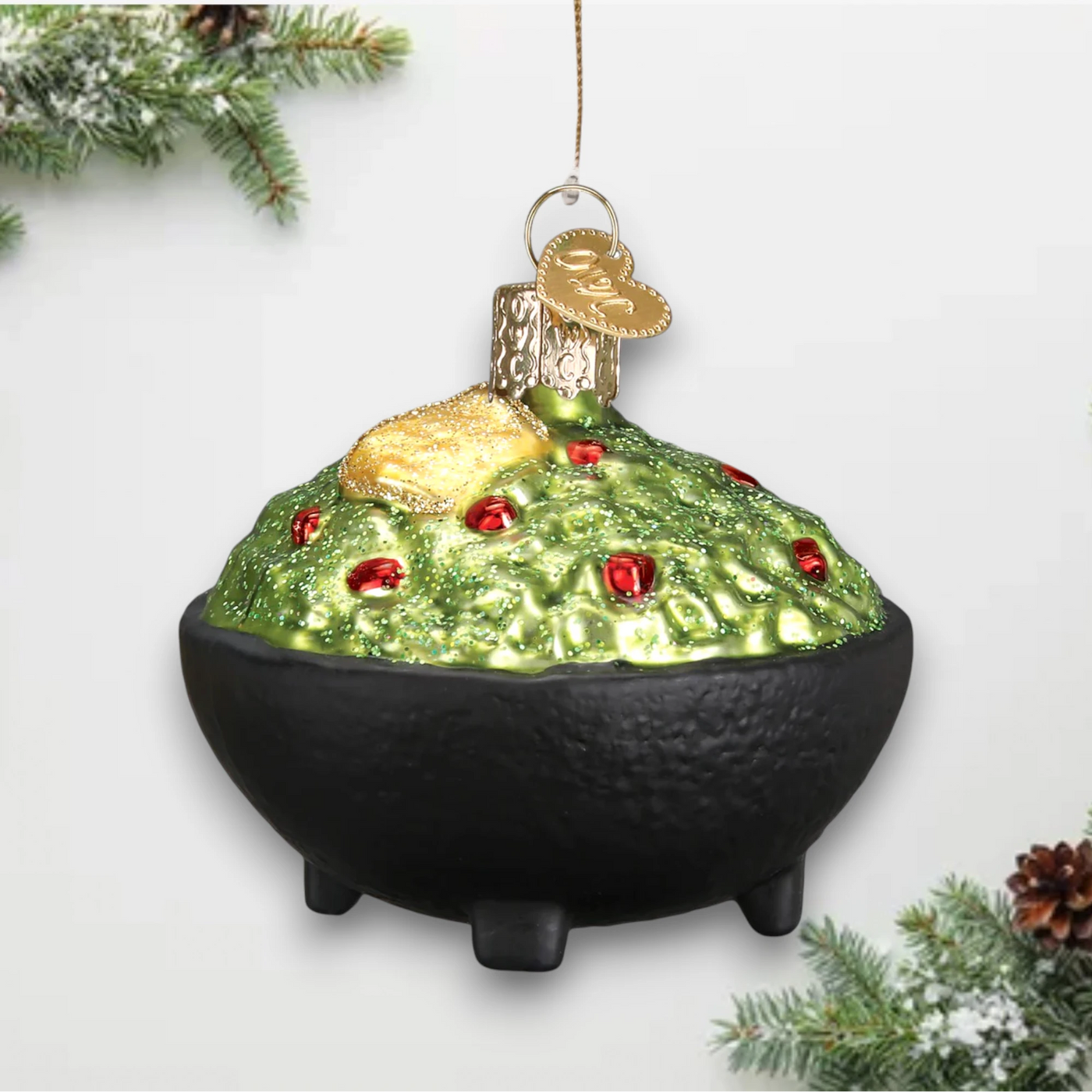 A glittery Christmas ornament depicting a bowl of guacamole with a golden tortilla chip on top, detailed with red accents and a black bowl base.