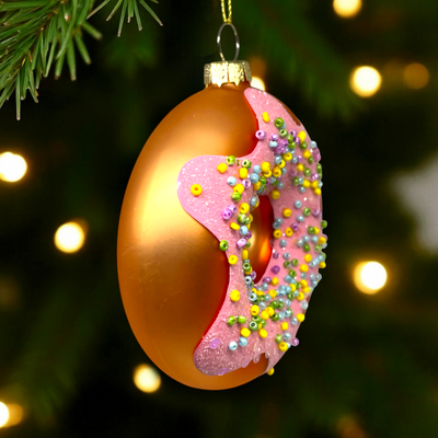 Decorative Christmas ornament shaped like a donut with pink icing and sprinkles, hanging on a tree.